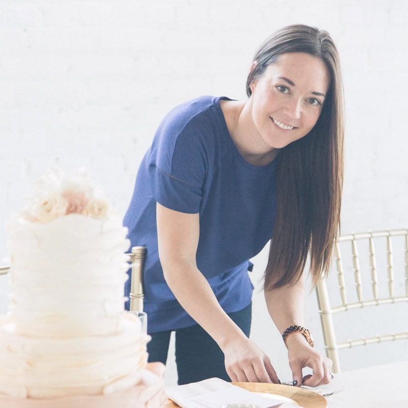 Bright smiling woman in casual blue attire preparing for a wedding event with a beautiful cake in the foreground at Melanie Parent Events, Winnipeg wedding and event planning specialists.