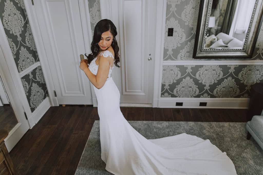 Elegant bride in a white wedding gown with lace details, standing indoors in a sophisticated setting with ornate wallpaper and a large mirror.
