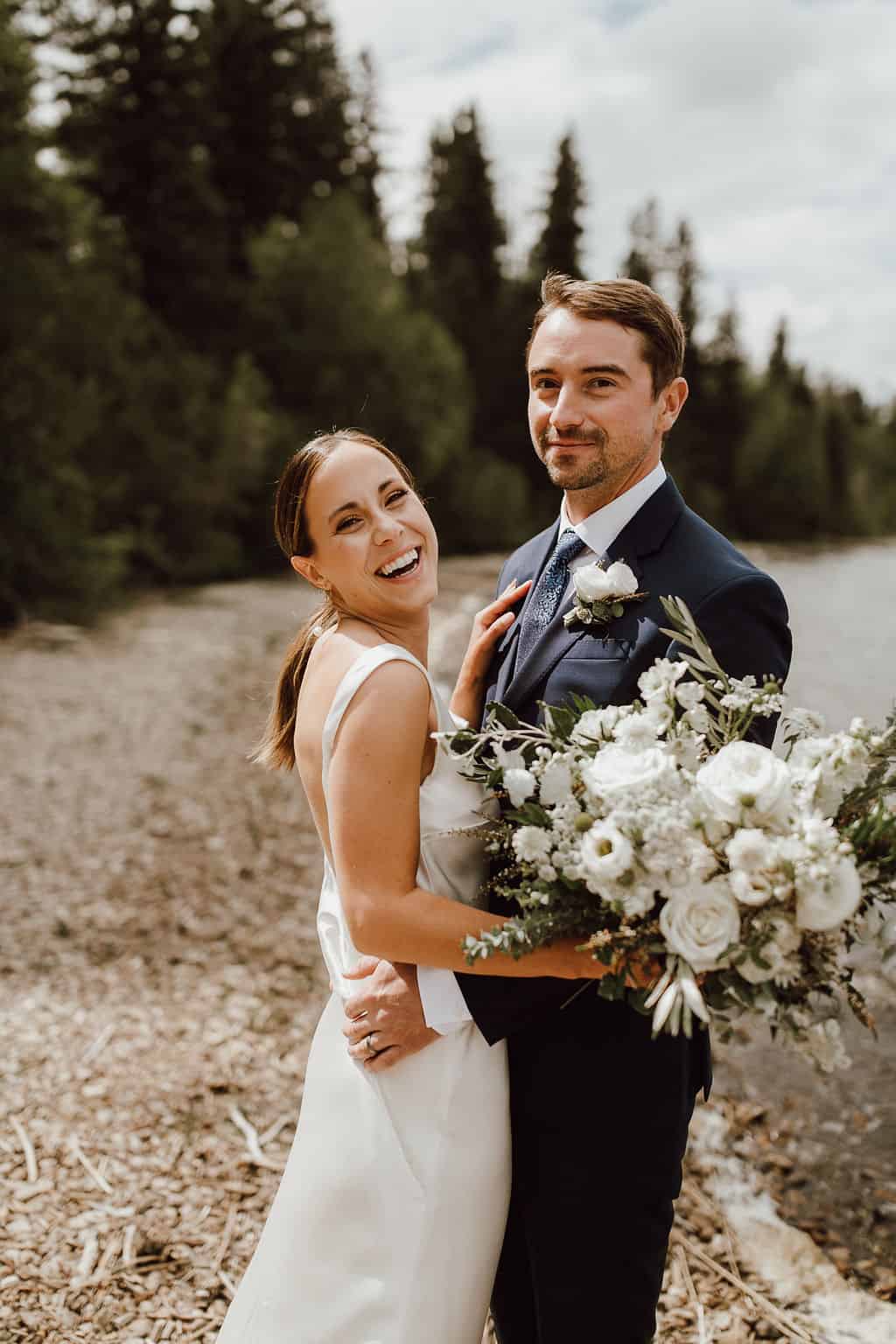 Elegant bride and groom smiling outdoors on wedding day with lush forest and river background, holding bouquet of white flowers.
