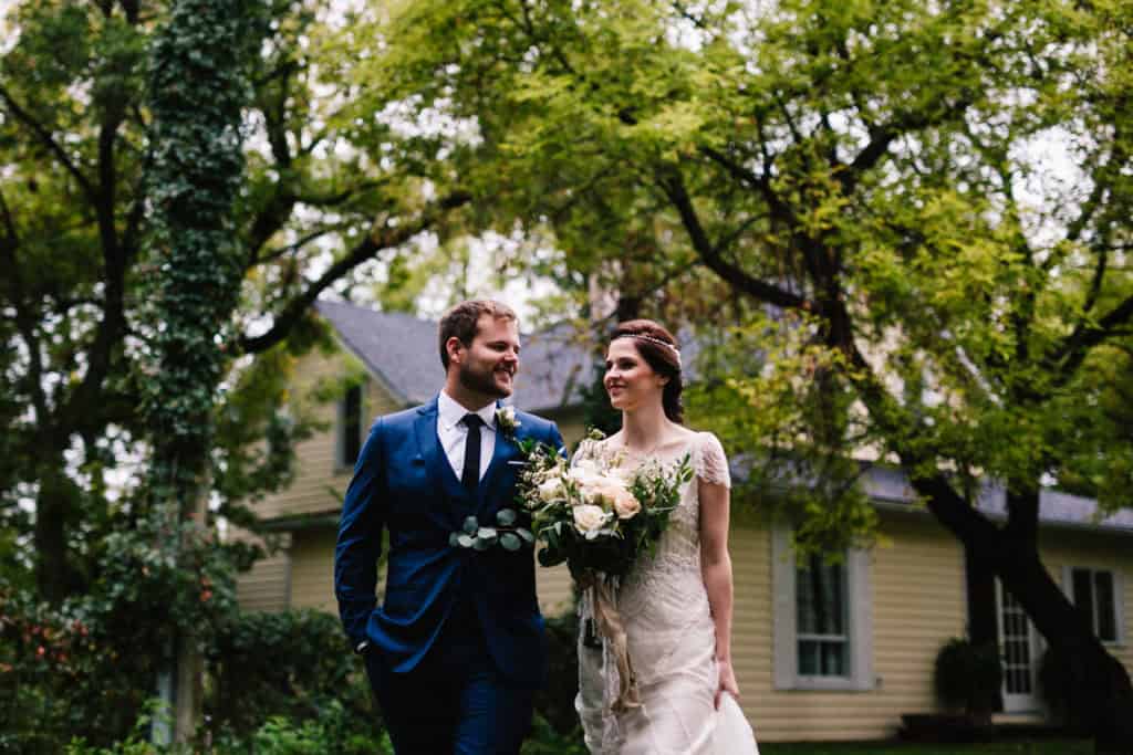 Elegant bride and groom walking outdoors at a wedding in Winnipeg, surrounded by lush green trees and a charming house, captured by Melanie Parent Events.