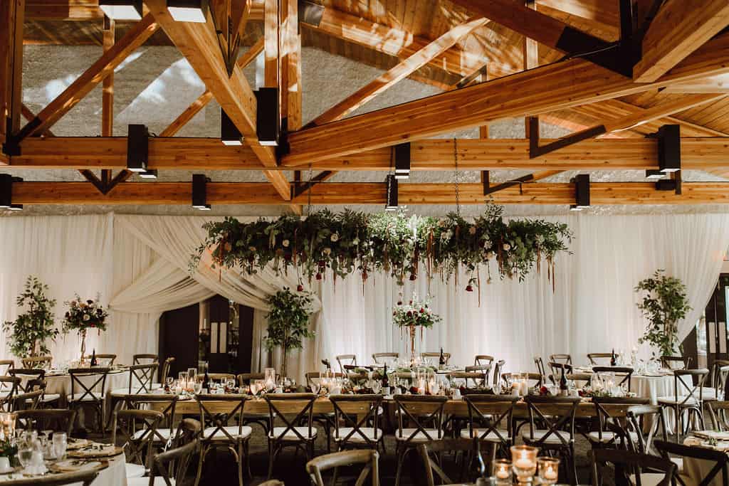 Elegant wedding reception decor with wooden ceiling beams, lush greenery, and floral centerpieces in a rustic yet sophisticated venue in Winnipeg.