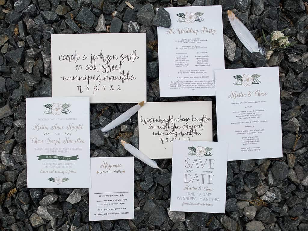 Elegant wedding stationery suite laid out on rocks, including save the date, invitations, response card, and addresses, featuring floral accents and stylish calligraphy for a Winnipeg wedding.