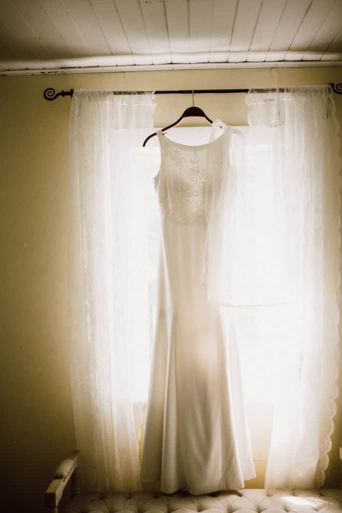 Elegant wedding dress hanging in front of sheer curtains, ready for bride on wedding day in Winnipeg.