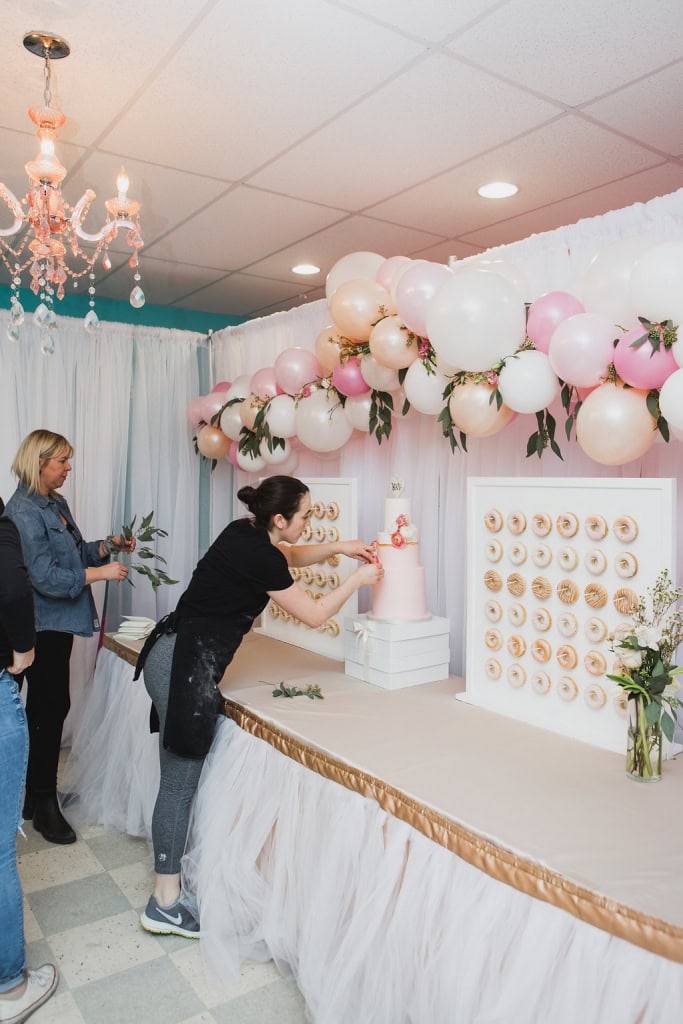 Pink and white balloon garland decorates a wedding cake table at Winnipeg event venue, with guests preparing to cut a pink fondant cake surrounded by balloon and donut wall displays.