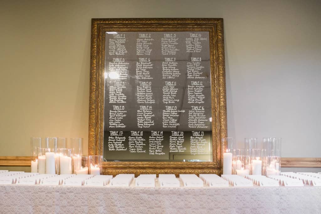 Handwritten wedding seating chart on a blackboard in an ornate gold frame with candles and white place cards on a table.