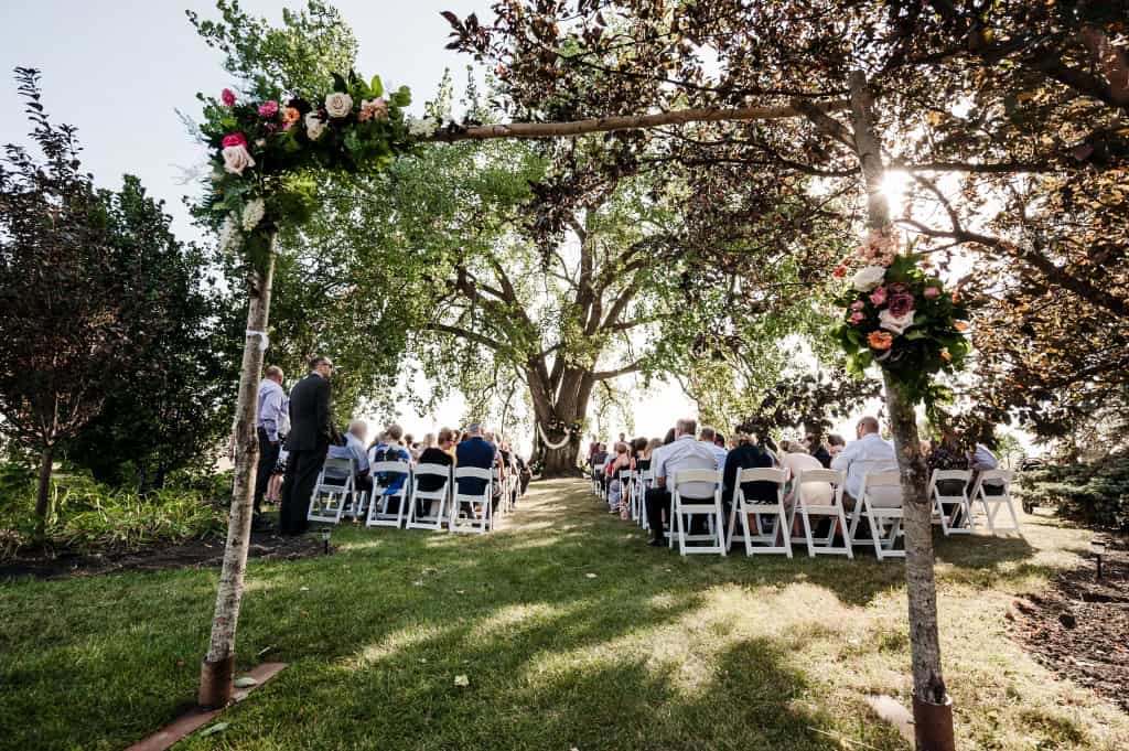 Beautiful outdoor wedding ceremony under blooming trees with guests seated on white chairs, sun shining through the branches.