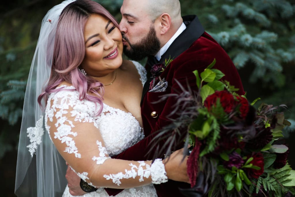 Vibrant wedding photo of a bride with lavender hair and a groom with a beard, sharing a joyful moment outdoors with lush greenery in the background.