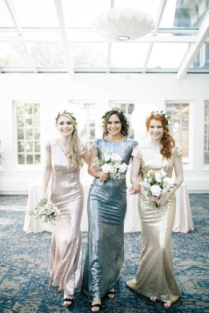 Elegant bridesmaids wearing shimmering metallic gowns, holding white floral bouquets, posing inside a bright, glass-ceiling event space in Winnipeg.