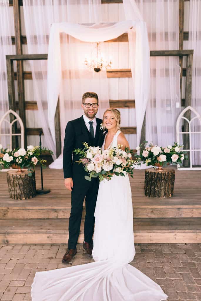 Elegant wedding couple in a rustic barn wedding setting with floral arrangements and draped fabric canopy in Winnipeg, captured by Melanie Parent Events.