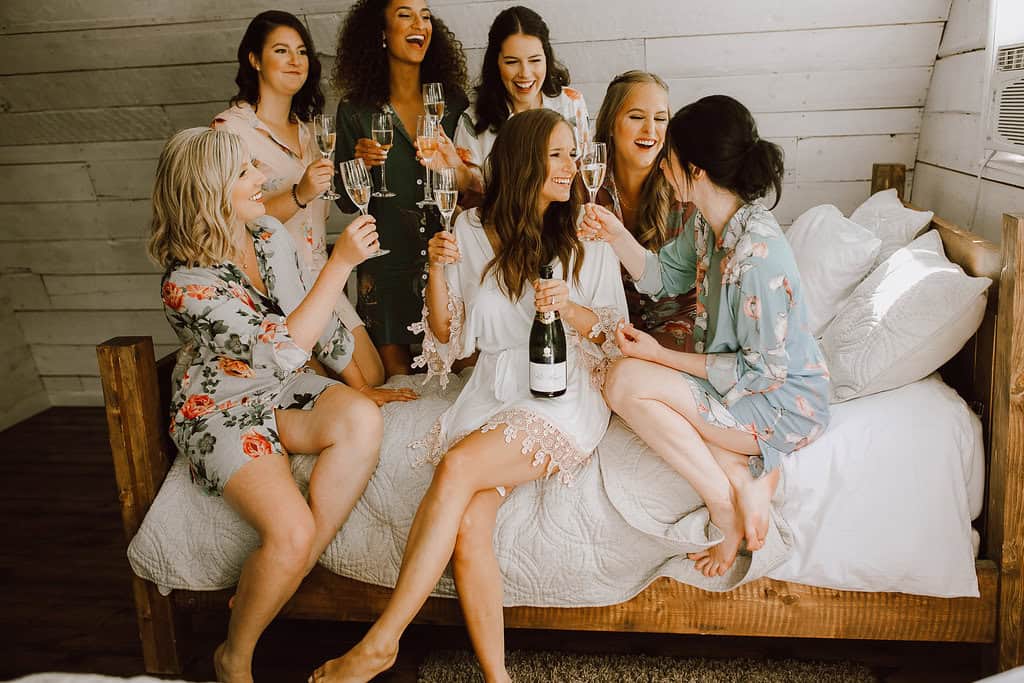 Six women in floral pajamas celebrating with champagne on a bed, enjoying a joyful moment, wedding or bridal party preparation, cozy indoor setting, friendly atmosphere.