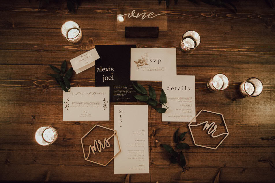 Elegant wedding invitation suite with greenery accents, candles, and "Mr" and "Mrs" wooden signs arranged on rustic wooden table for wedding decor.
