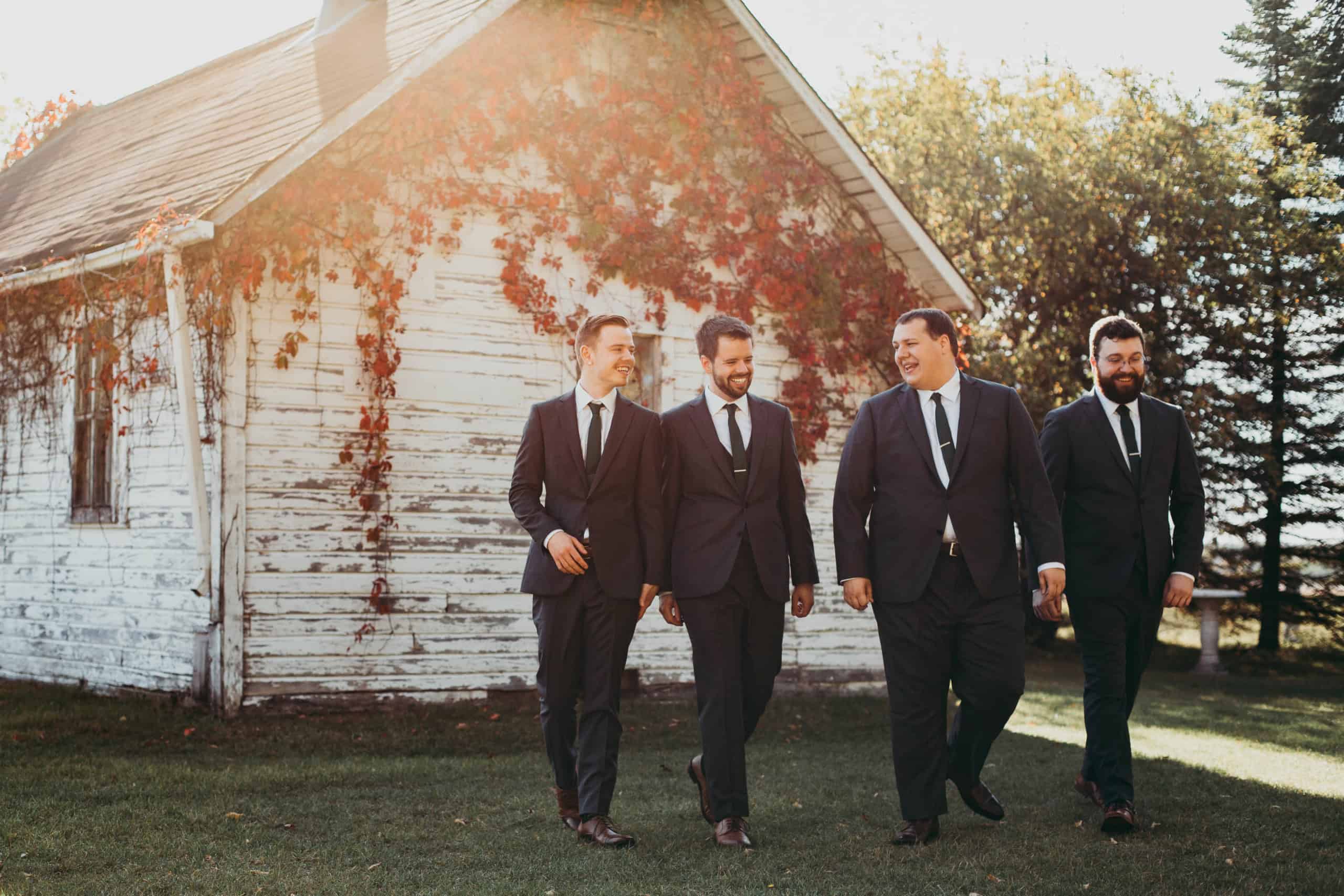 Elegant men in black suits walking outside near a rustic white barn with autumn foliage in the background.