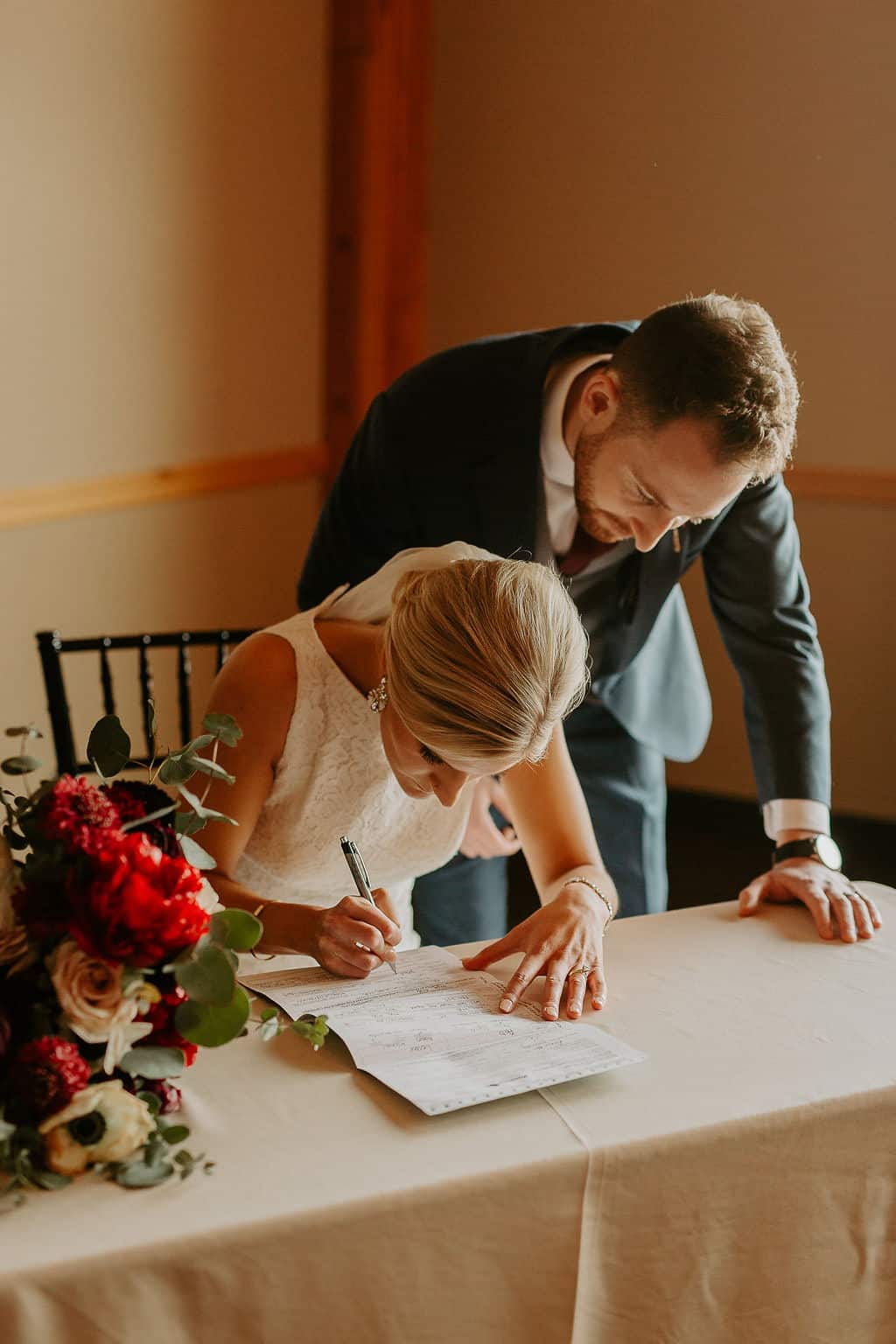 Elegant bride signing wedding registry while groom assists at a wedding signing table.