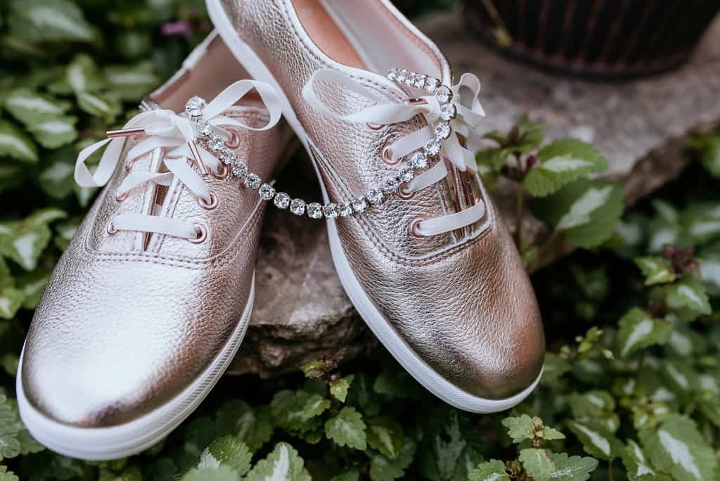 Shiny rose gold sneakers with white laces and a diamond-studded bracelet sitting on green foliage. Perfect for wedding or floral event accessories.