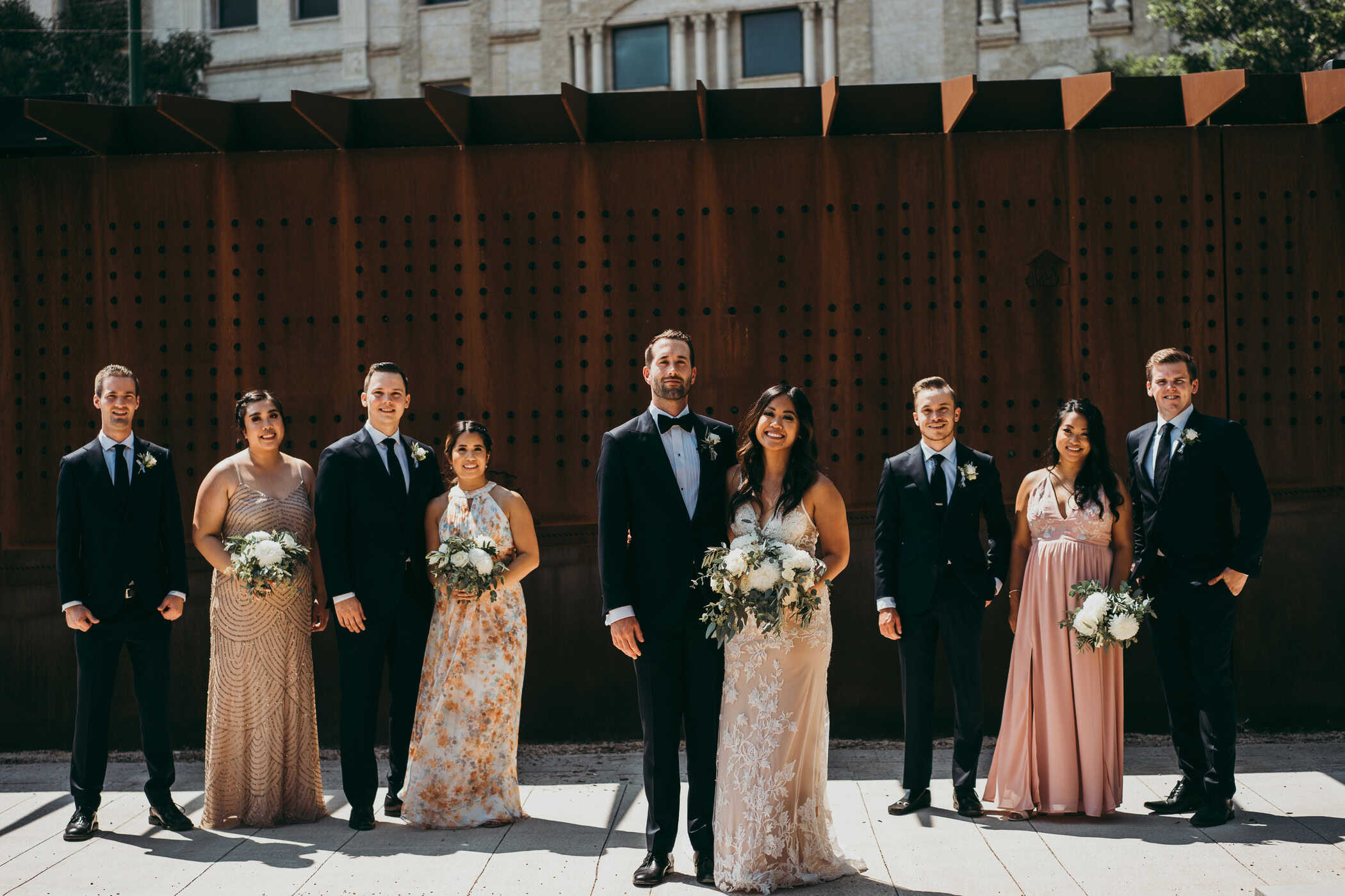 Elegant wedding party in formal attire with bouquets, standing outdoors against a stylish wooden backdrop in Winnipeg, celebrating a wedding at Melanie Parent Events.