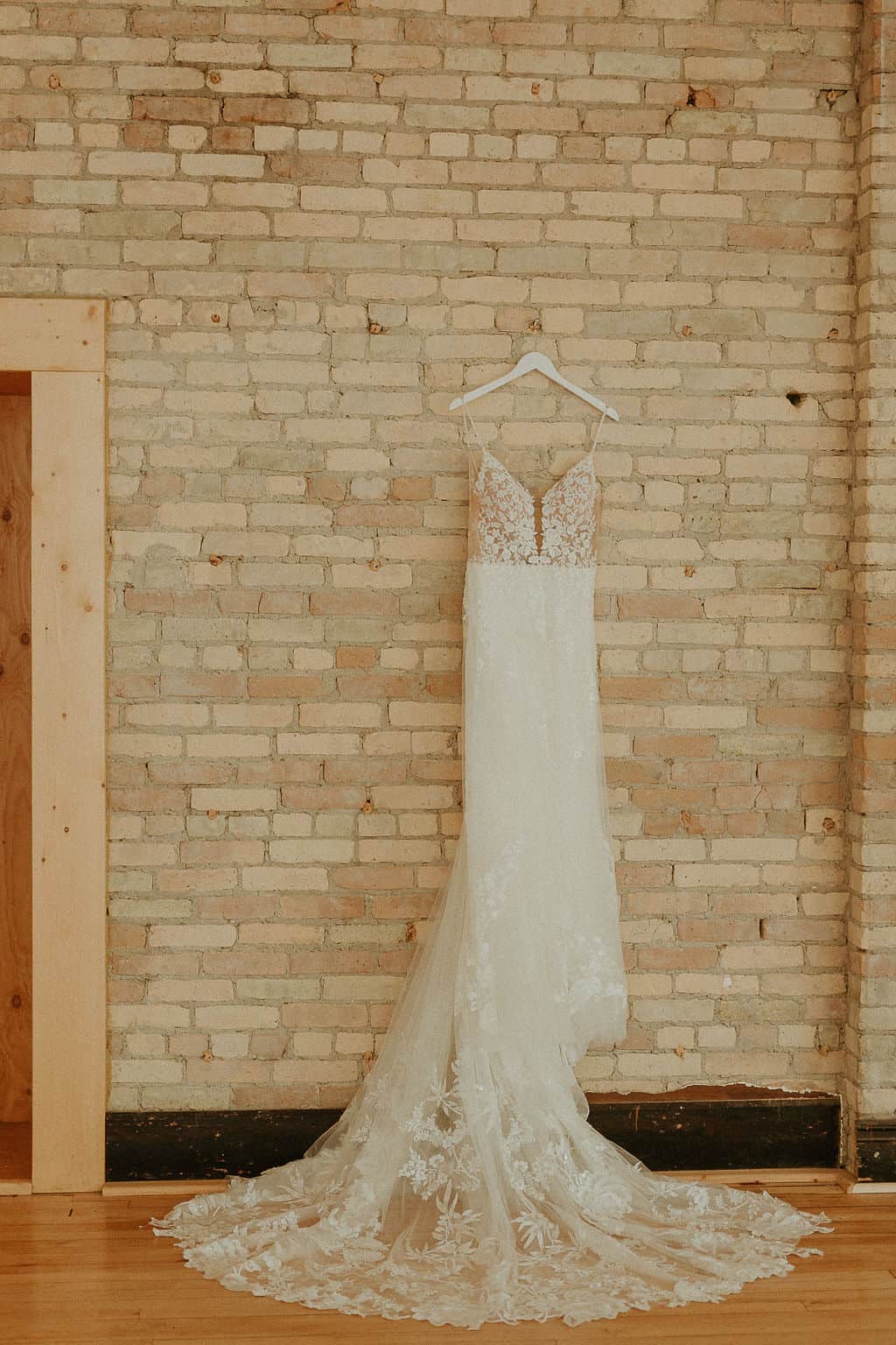 Elegant lace wedding dress hanging on a white hanger against a brick wall.