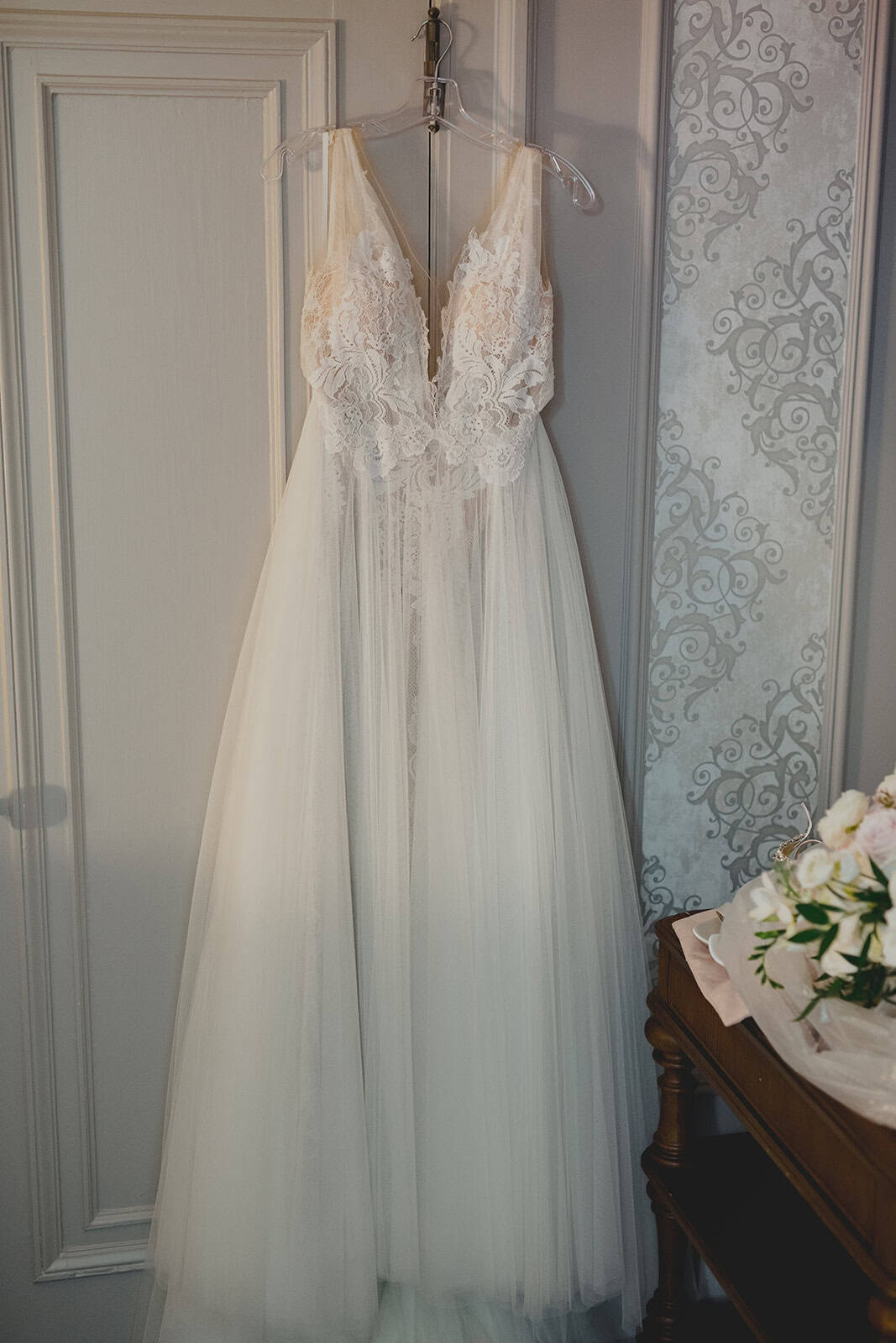 Elegant white wedding dress with lace bodice and flowing tulle skirt hanging on a clear hanger in a bridal suite.