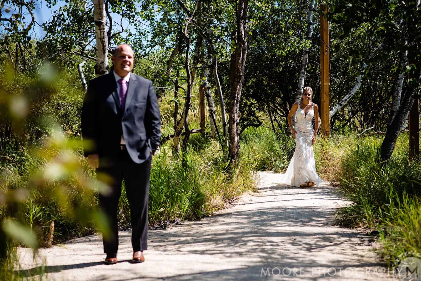 Blurry image of a man in a suit standing on a trail with a bride approaching in a white wedding gown among trees and greenery.