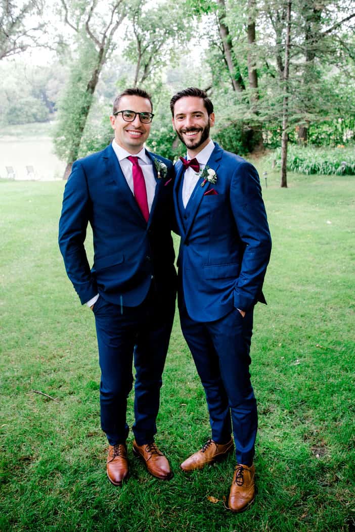 Elegant groom and groomsman smiling in a park during wedding celebration, both dressed in sharp navy suits with burgundy accents.