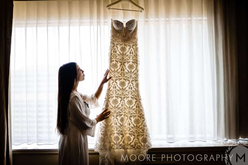 Elegant wedding dress hanging in front of sheer curtains with bride admiring the gown.