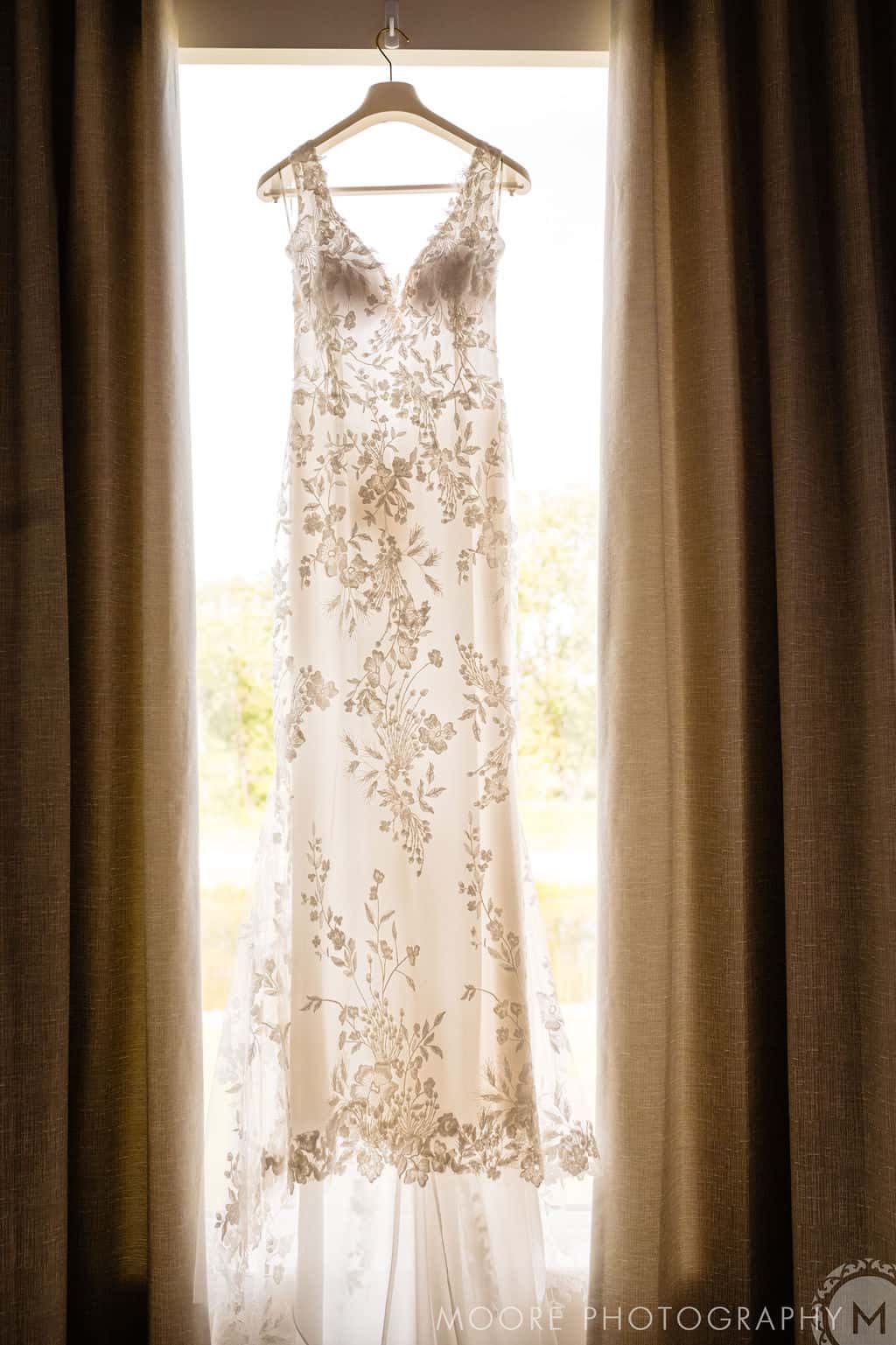 Elegant wedding dress with floral lace details hanging in front of a backlit window, ready for a bride's special day at Melanie Parent Events in Winnipeg.