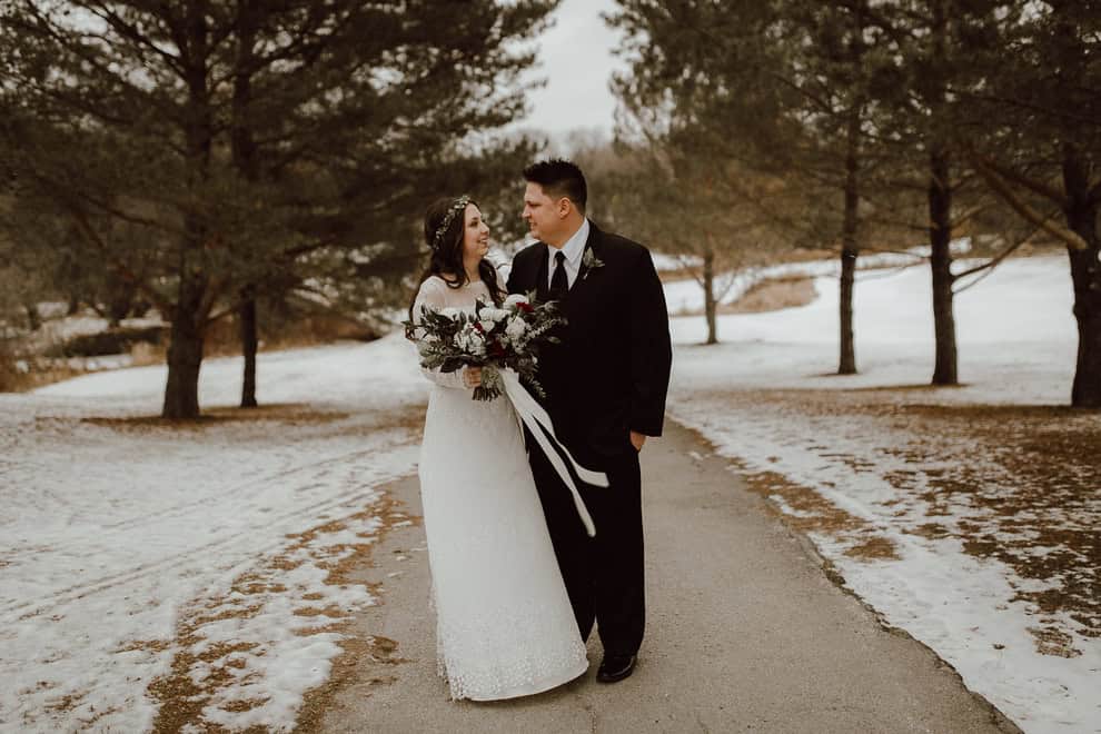 Elegant wedding couple walking outdoors on a winter day with snow-covered ground and trees in the background, celebrating love and marriage.