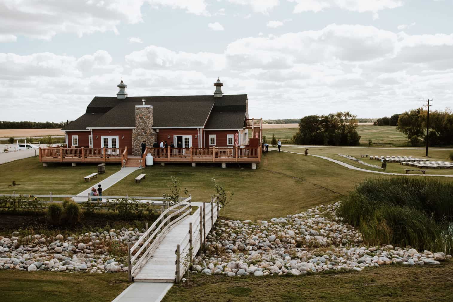 Charming red barn-style event venue with a wooden deck, surrounded by open fields, landscaped lawn, and a pond with a white footbridge, ideal for outdoor weddings and celebrations.