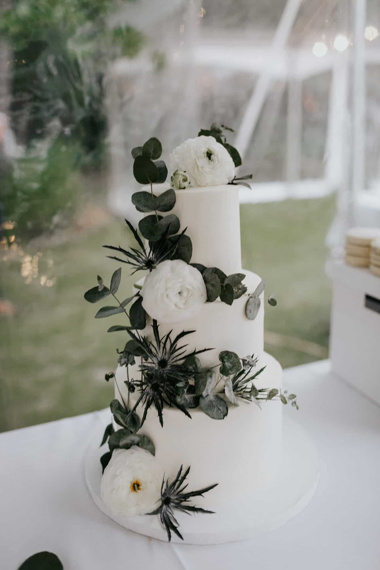 Elegant white wedding cake decorated with white ranunculus flowers and dark greenery, displayed on a white table at an outdoor wedding reception.