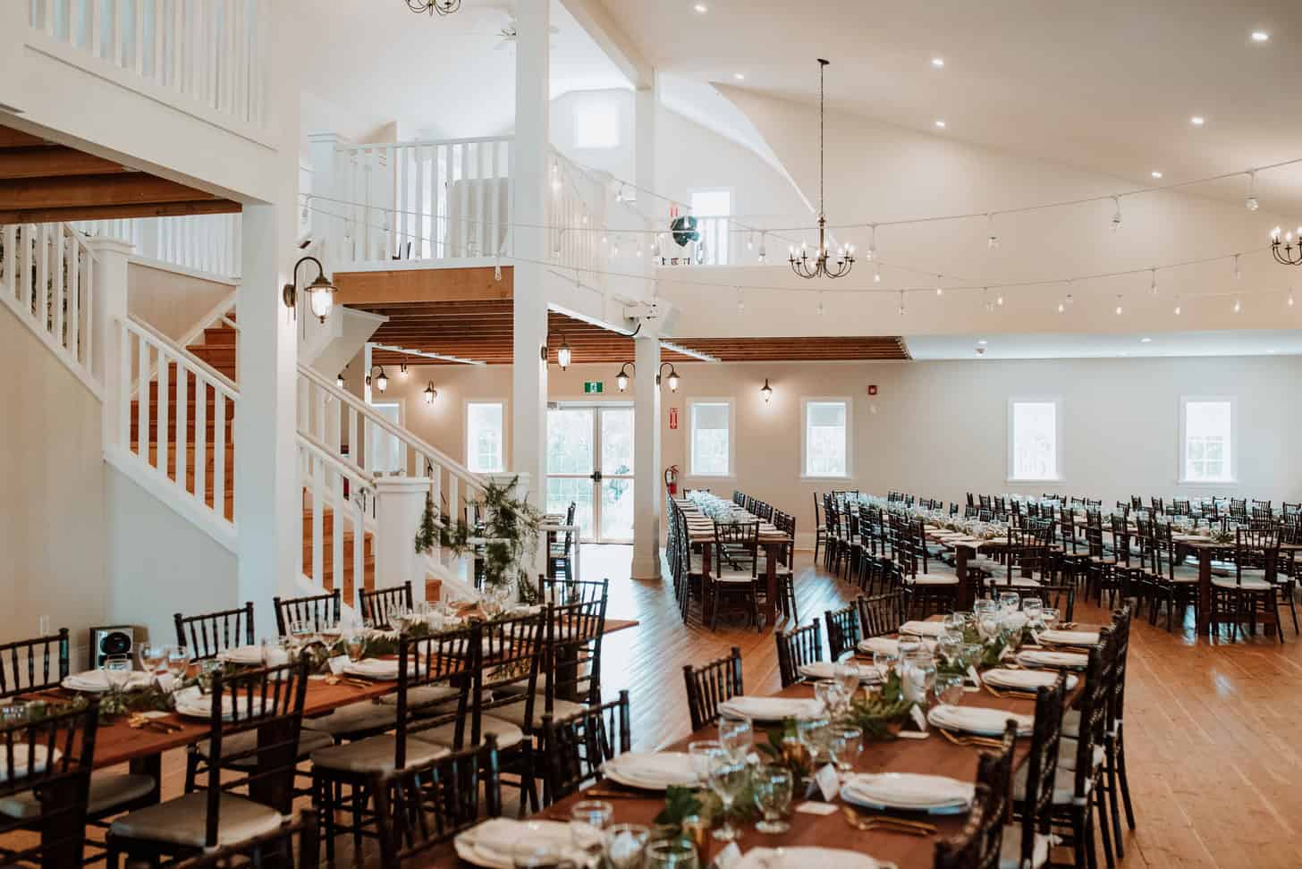 Elegant wedding reception setup in a spacious, well-lit venue with long wooden tables, black chairs, tasteful table settings, and delicate string lighting.