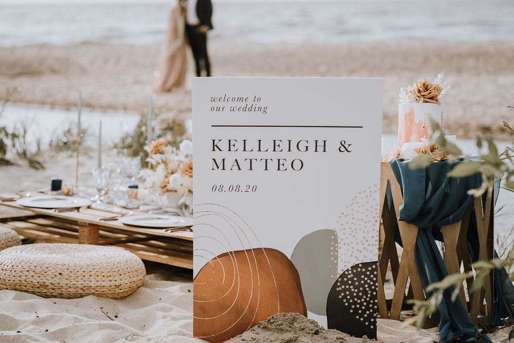 Bright beach wedding setup with a personalized wedding sign, elegant table settings, and rustic accents on the sand, creating a romantic seaside celebration vibe.
