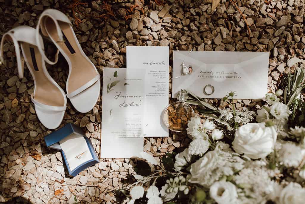 Elegant white wedding shoes, jewelry, wedding invitation, bouquet, and wedding rings displayed on a gravel surface for a wedding celebration.