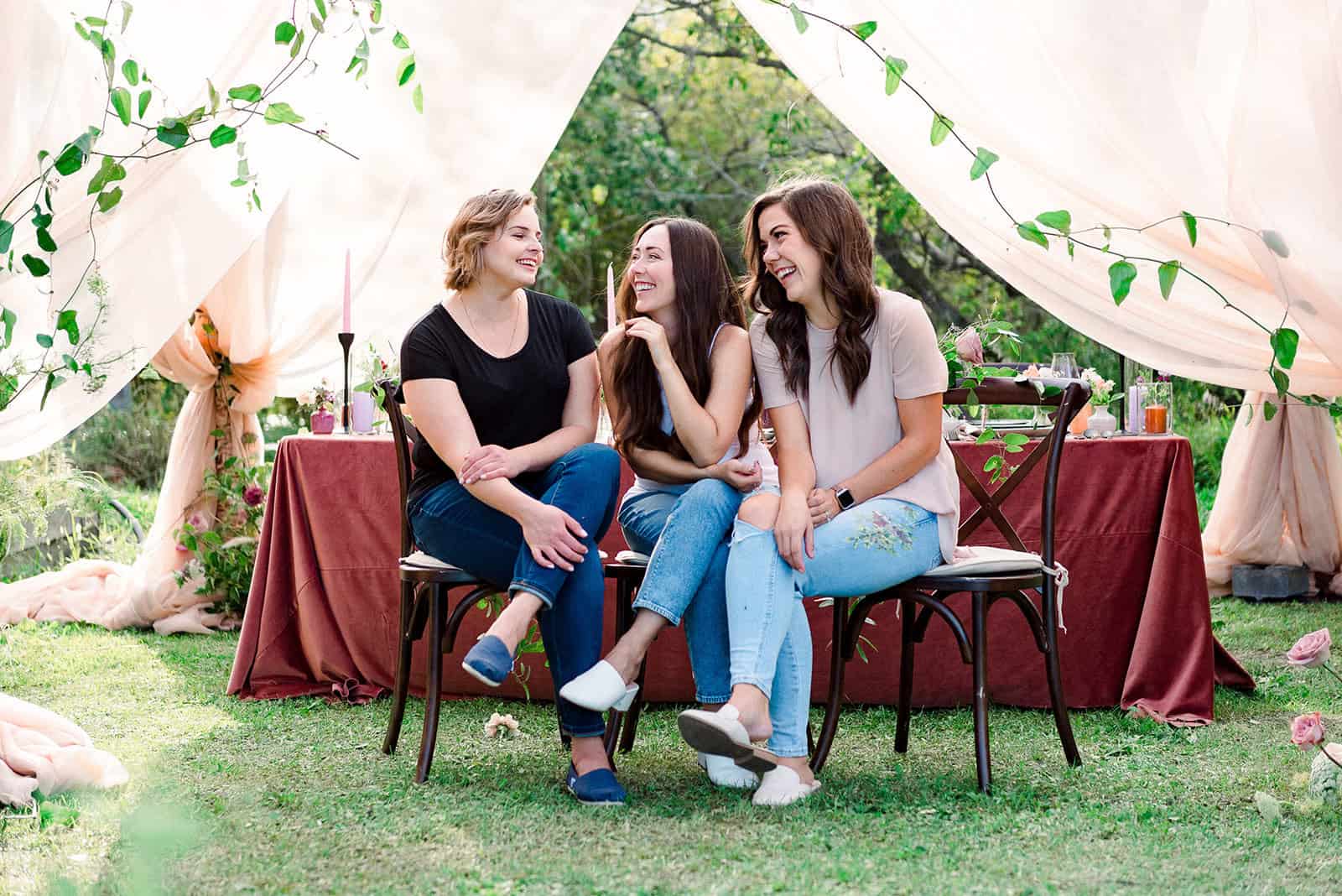 Vibrant outdoor wedding reception with three women sitting on chairs under a decorated tent, surrounded by lush greenery, floral accents, and elegant table settings, enjoying a joyful moment.