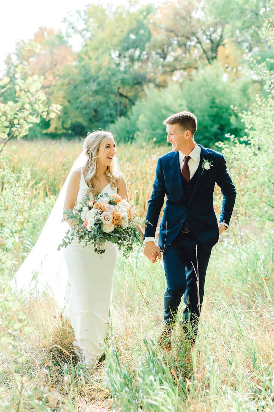 Elegant bride and groom holding hands in a scenic outdoor setting, celebrating their wedding day in a lush, green landscape with vibrant foliage and natural sunlight.