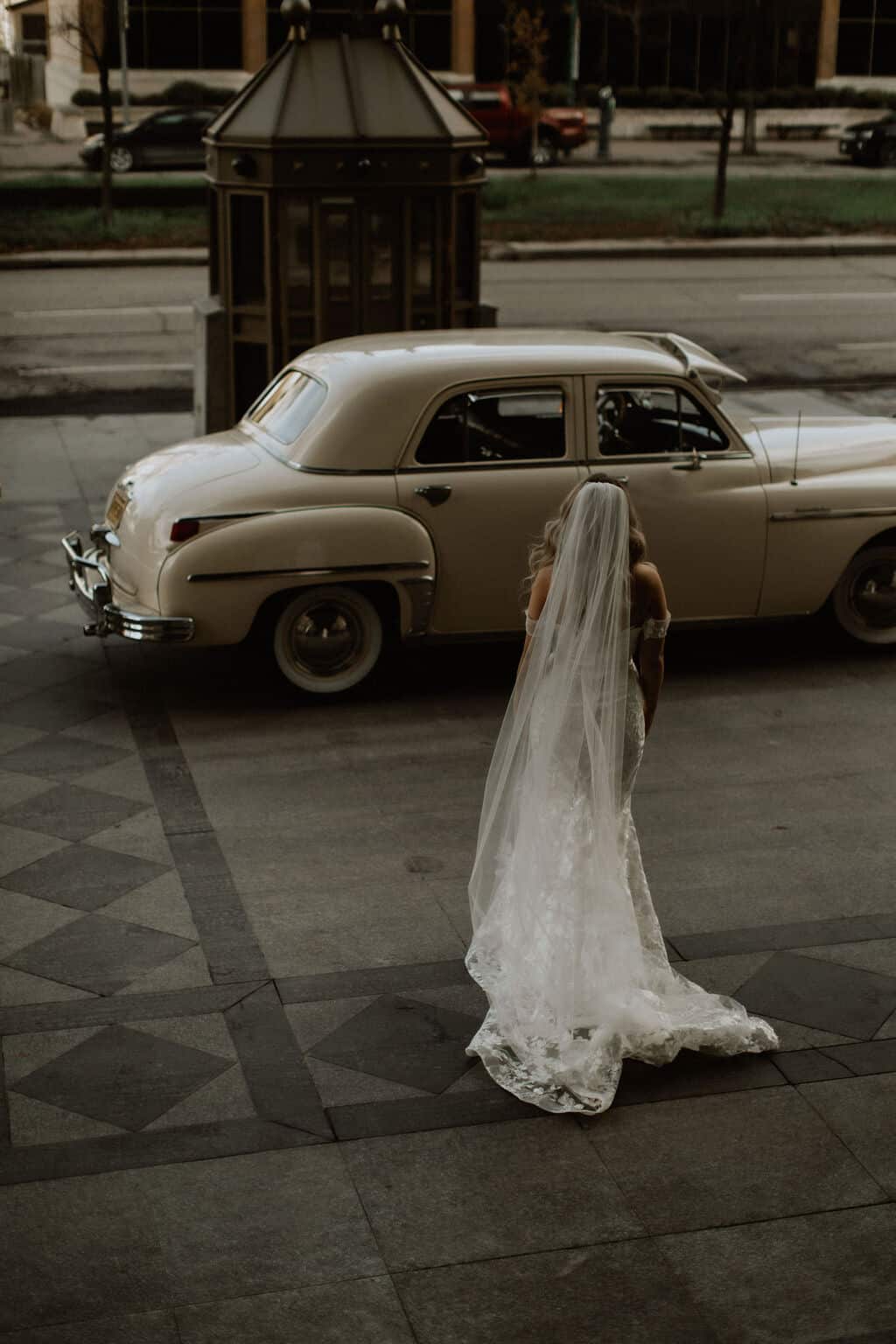 Vintage cream-colored car parked on city sidewalk with a bride in a white wedding gown and veil standing next to it, outdoor urban scene, elegant wedding transportation shot.