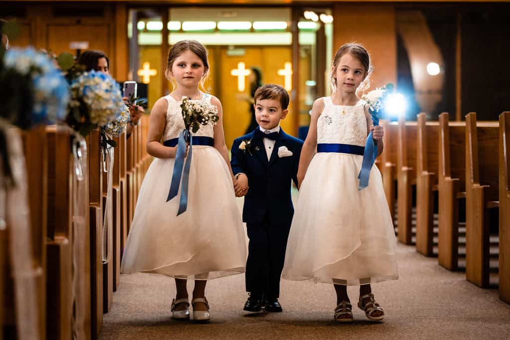 Cute young ring bearer and flower girls walking down church aisle during wedding ceremony, children in formal attire holding flowers and smiles.