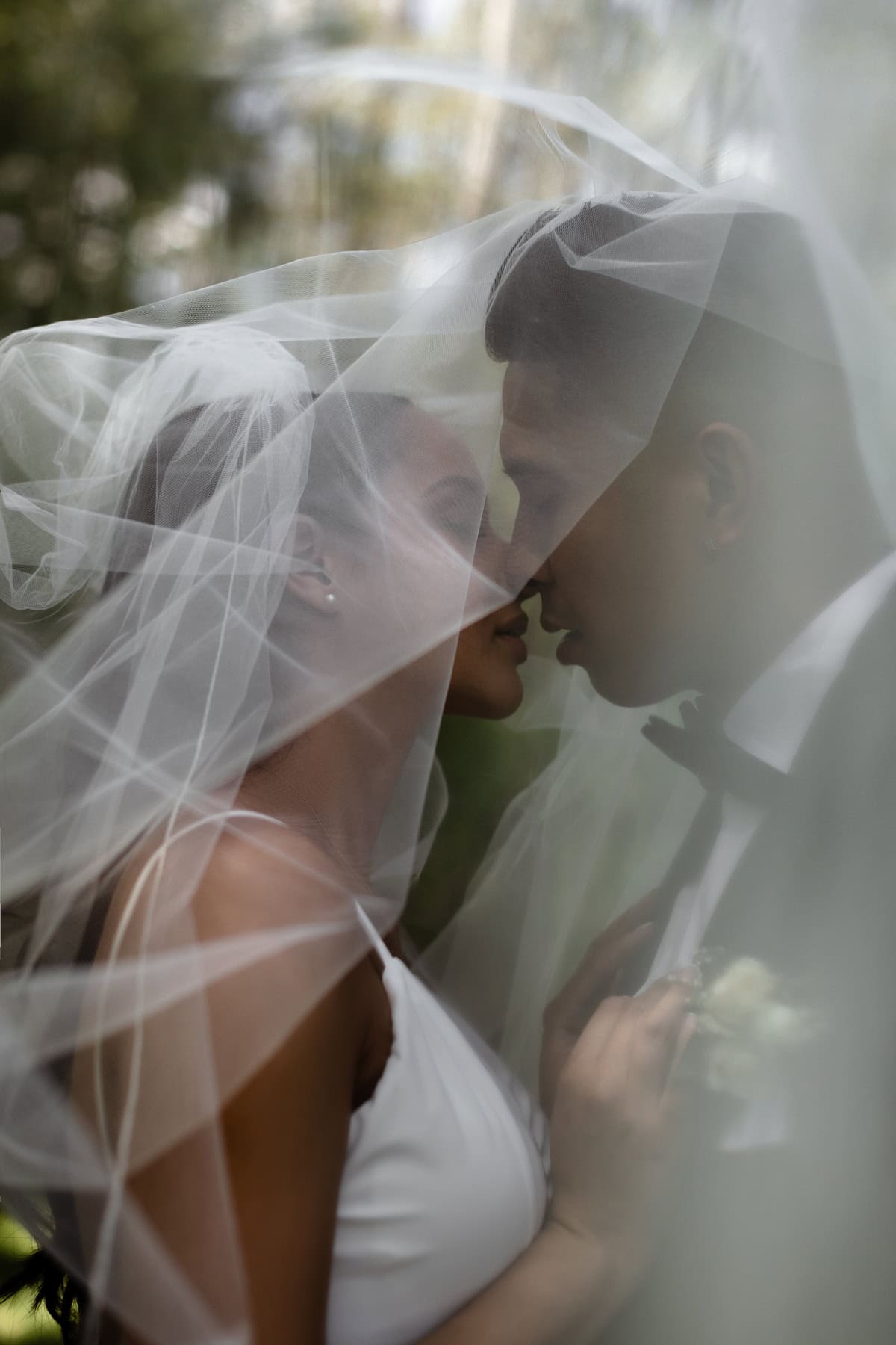 Elegant couple sharing an intimate moment under a wedding veil outdoors, with a soft natural background highlighting the romance and beauty of a wedding day.