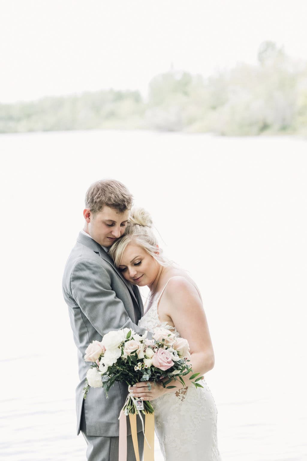 Elegant wedding couple embracing by the water with a bouquet of pink and white roses, capturing a romantic moment during their outdoor wedding celebration.