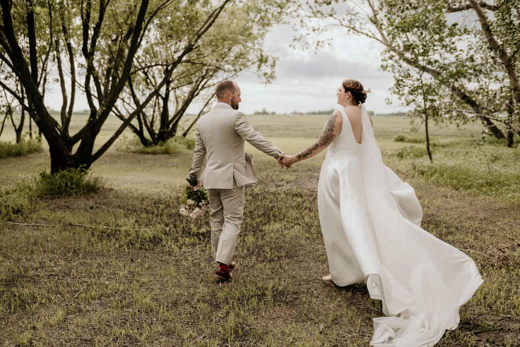 Romantic wedding couple holding hands outdoors in a natural grassy setting with trees, showcasing a beautiful wedding ceremony scene.