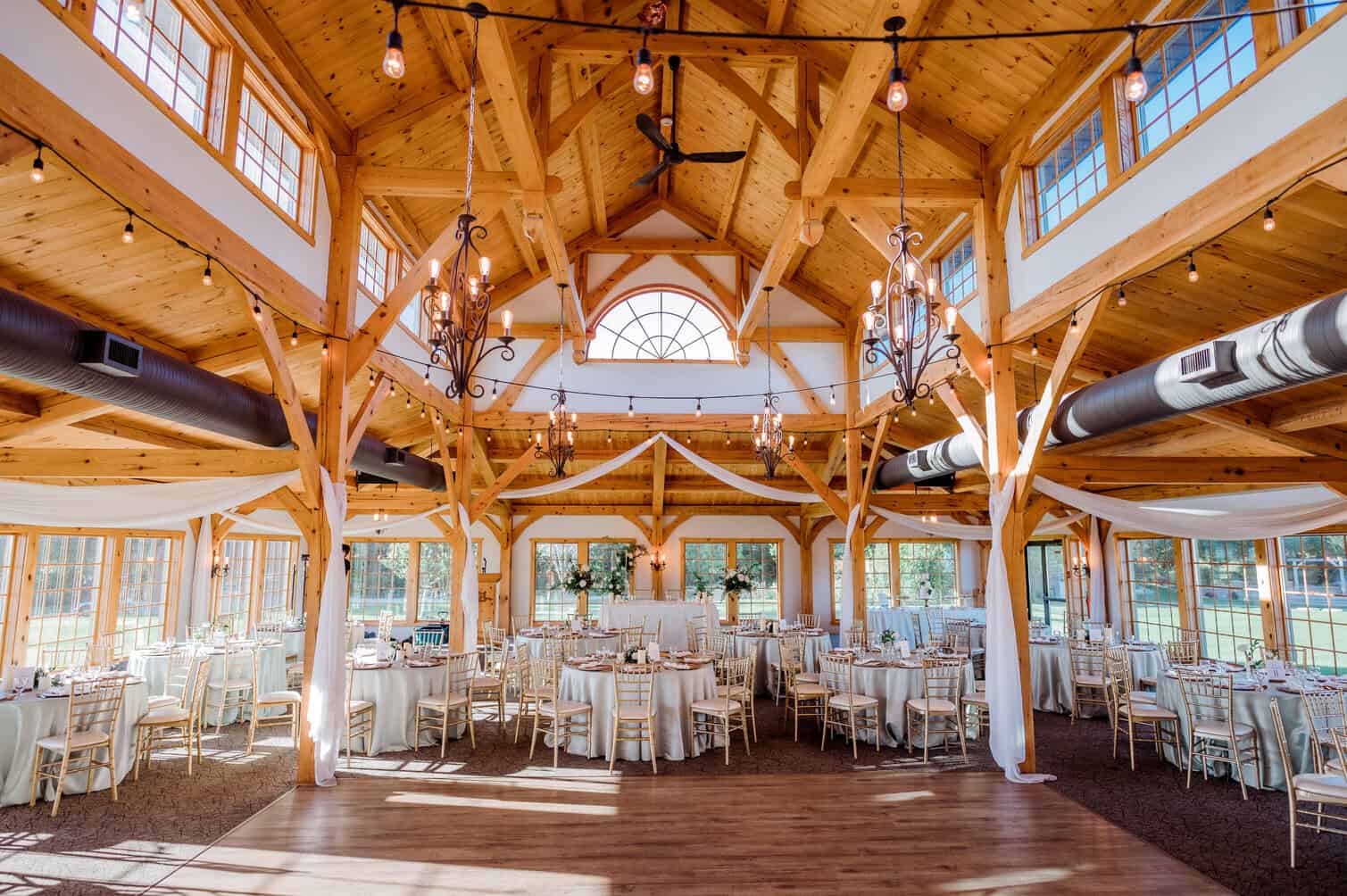 Elegant wooden wedding reception venue with high vaulted ceilings, chandeliers, string lights, and large windows in Winnipeg. Perfect for weddings and special events.