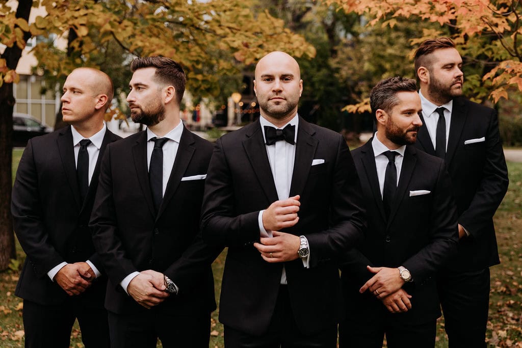 Elegant groom and groomsmen in black suits and tuxedos at an outdoor fall wedding, standing beneath colorful autumn trees in Winnipeg.