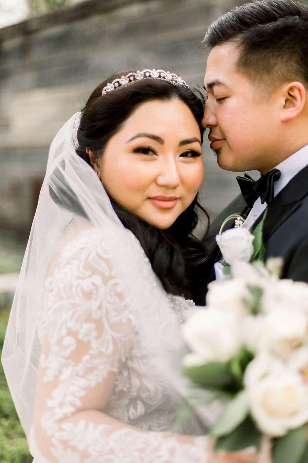 Elegant wedding portrait of a bride and groom, showcasing the bride's intricate lace gown and tiara, with the groom in a black tuxedo, capturing a romantic moment outdoors.