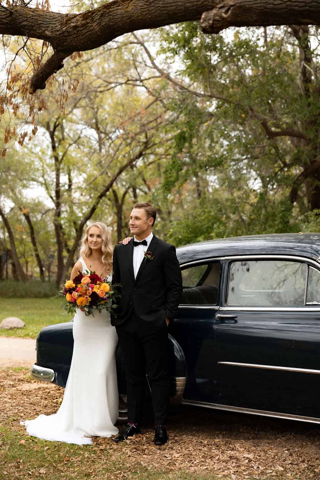 Beautiful wedding couple standing next to a vintage car in a lush, green outdoor setting with trees and foliage.