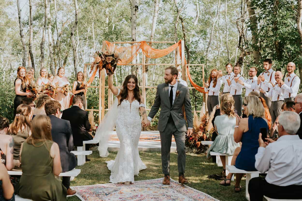 Bright outdoor wedding ceremony with bride and groom celebrating their marriage in a forest setting. Elegant floral arrangements, guests, and wedding party in formal attire.