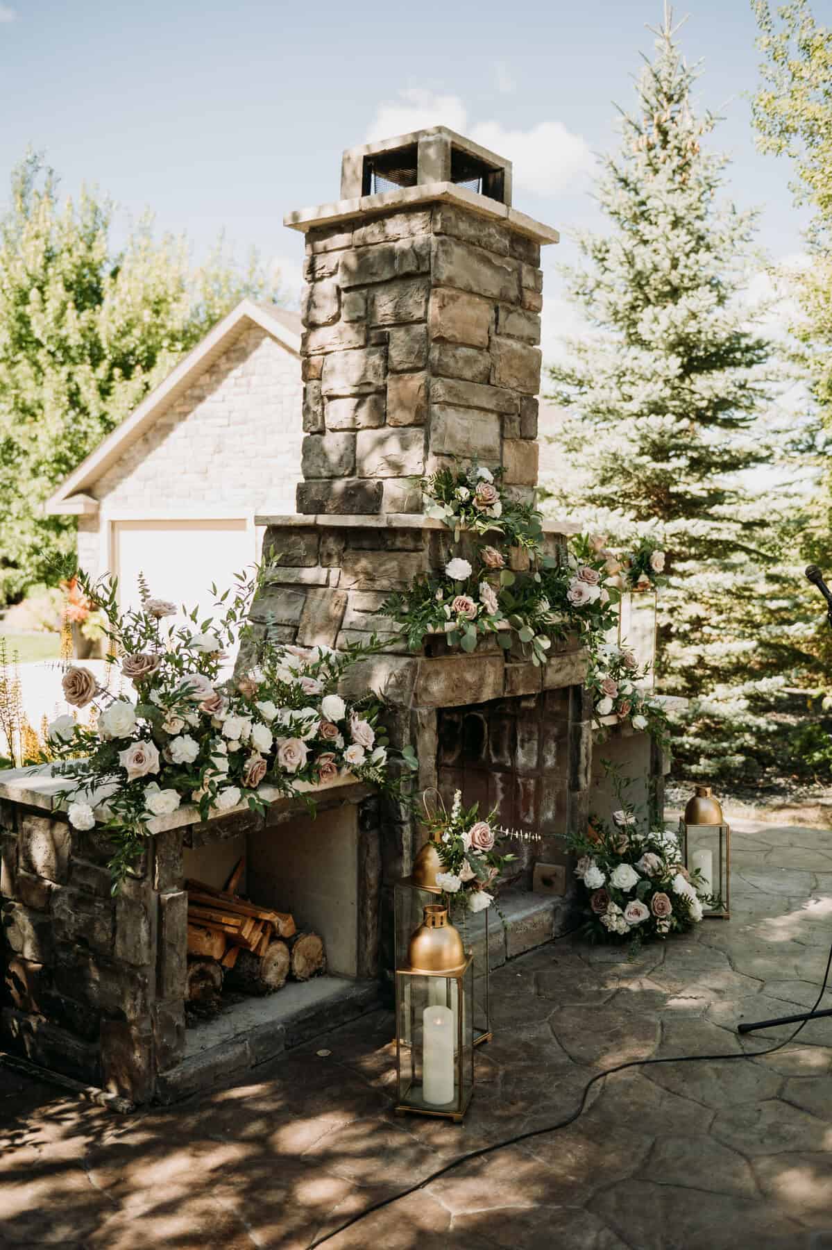 Elegant outdoor fireplace decorated with white and blush pink flowers, greenery, and lanterns for wedding or event decor in Winnipeg.