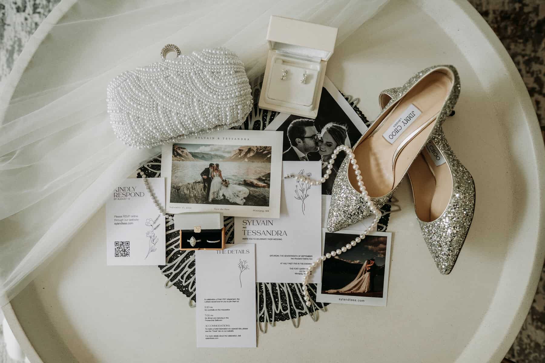 Pearl clutch and jewelry for wedding accessories, silver glitter high heels, wedding invitation set, and photos of a bride and groom on a white table.