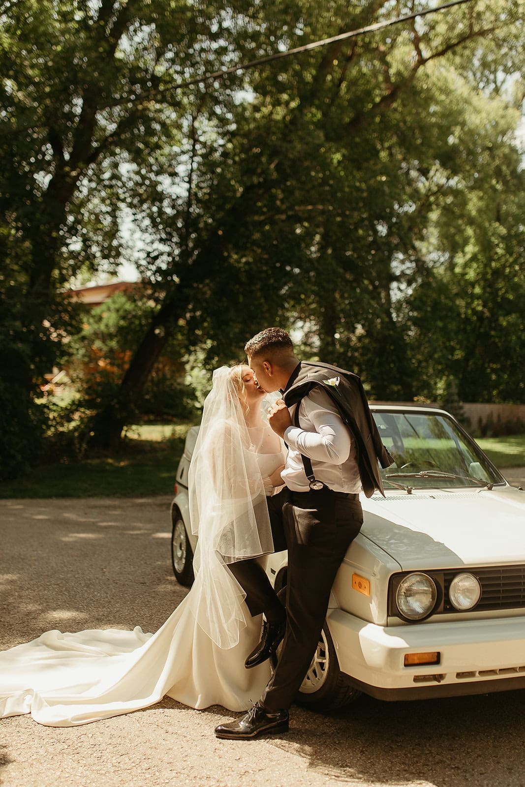 Elegant bride and groom sharing a kiss outdoors next to a classic white car during their wedding day in Winnipeg. Beautiful natural setting with lush green trees providing a picturesque backdrop.