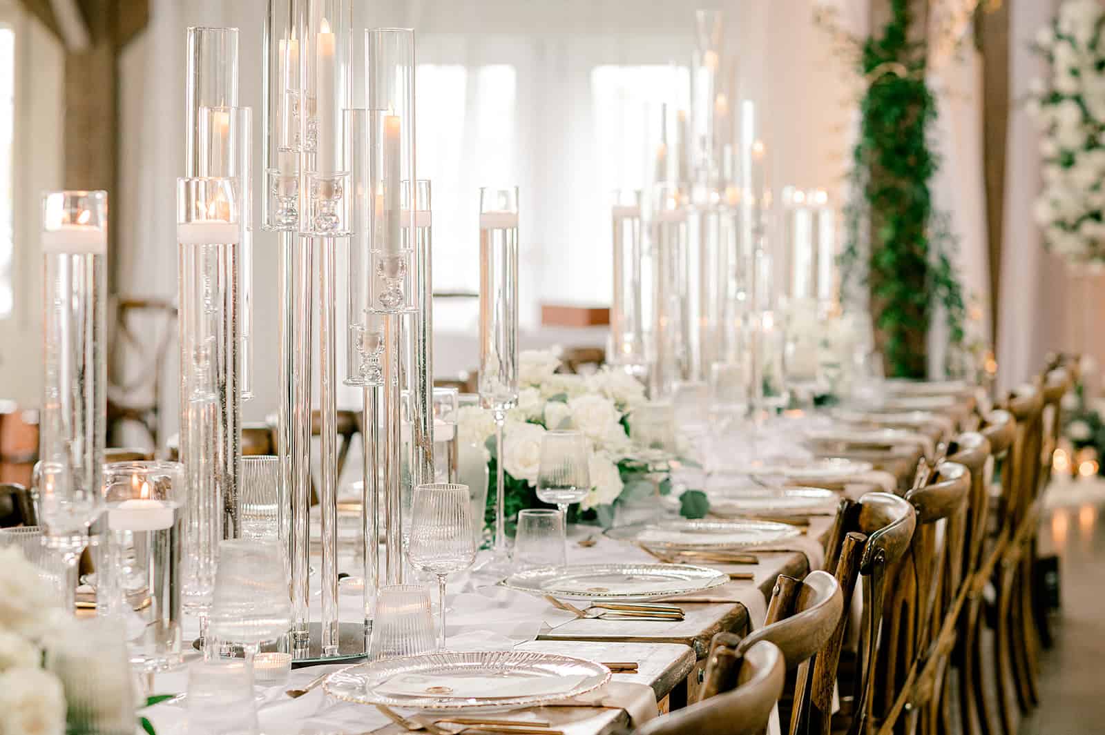 Elegant wedding reception table with tall glass candle holders, white floral centerpieces, and gold accents, set in a bright, sophisticated venue with natural light.