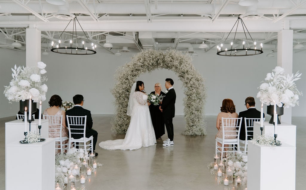 Elegant wedding ceremony setup with white floral arch, bridesmaids, groomsmen, and couple exchanging vows in a modern indoor venue with minimalist decor.