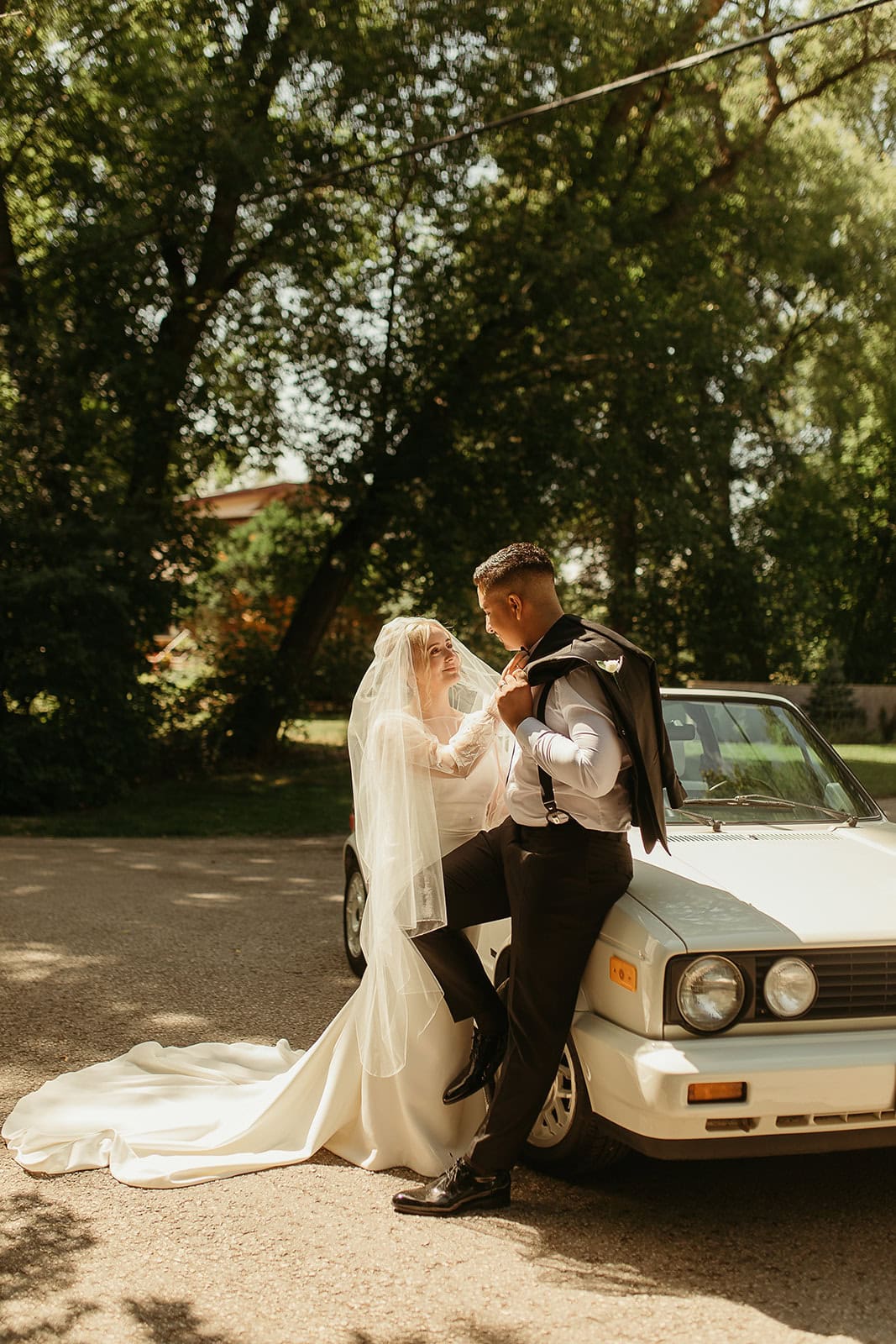 Elegant bride and groom in wedding attire leaning against a vintage car outdoors with lush green trees in the background, capturing a romantic moment on their wedding day.