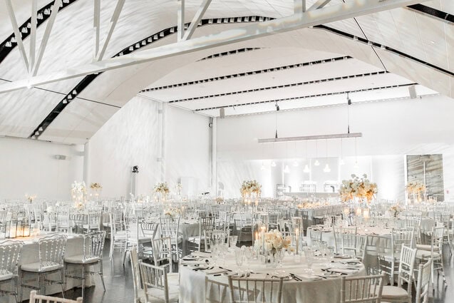 Elegant wedding reception setup with white chairs and floral centerpieces in a modern, airy venue.